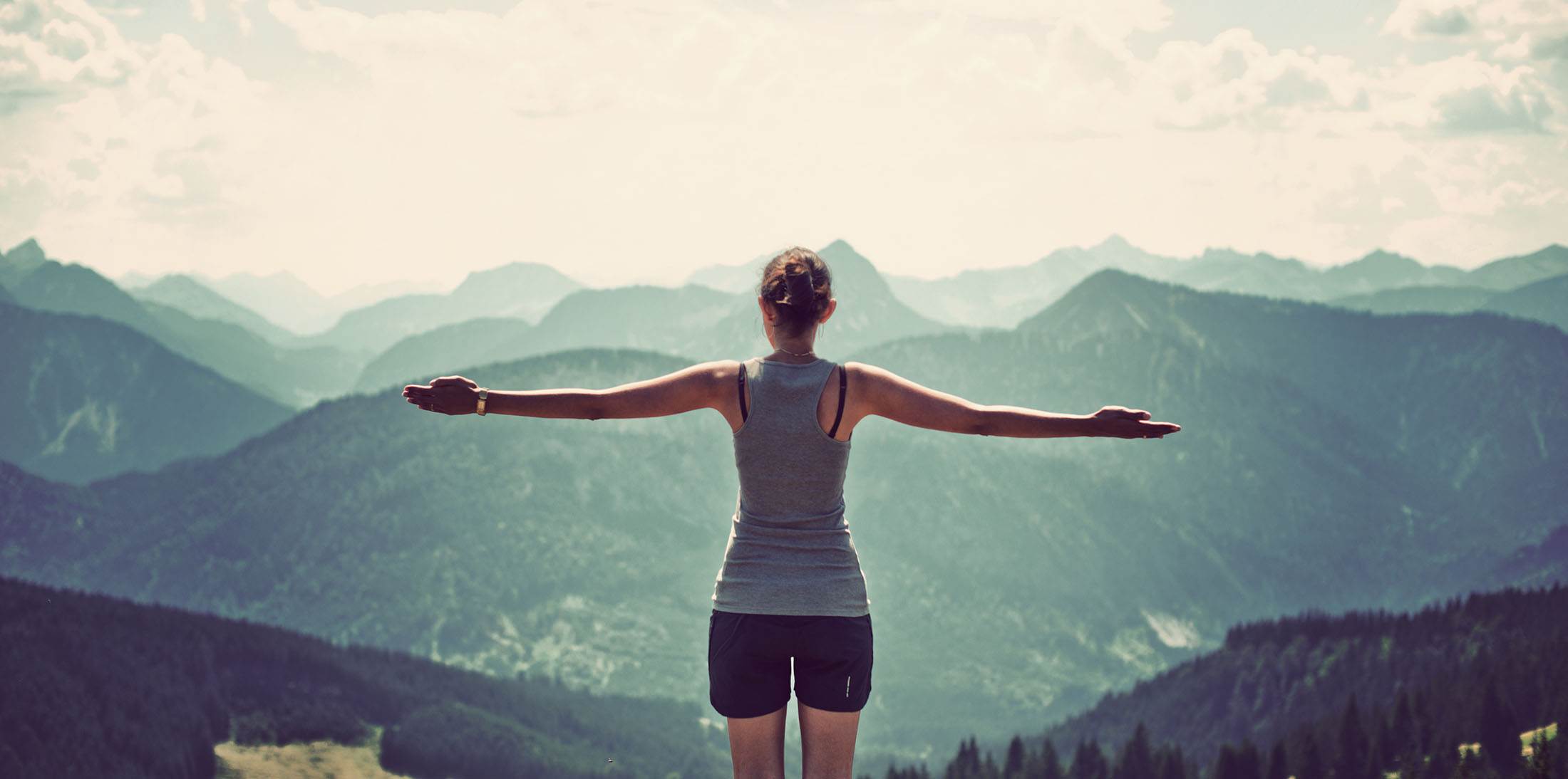 Woman celebrating nature and reaching the summit of a high mountain as she stands with her back to the camera and arms extended looking out over mountain ranges and valleys in a panoramic landscape